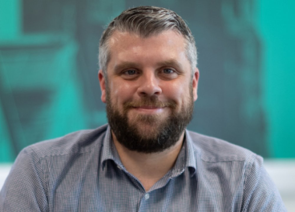 A person wearing a grey button‑up shirt sits in front of a teal backdrop featuring architectural illustrations and a large clock face.
