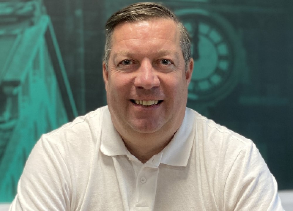 A person wearing a light-coloured polo shirt sits in front of a teal backdrop featuring architectural illustrations and a large clock face.
