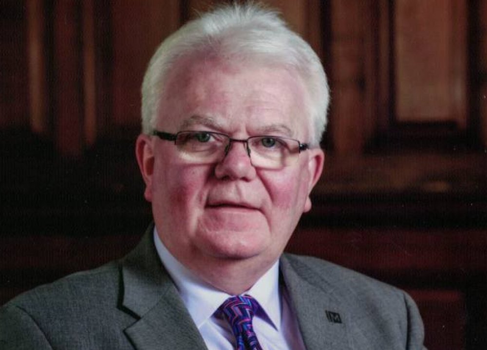 A person with short white hair is seated indoors wearing a grey suit jacket, white shirt, and a patterned purple tie. The background features dark wooden panelling, giving the scene a formal, traditional setting.
