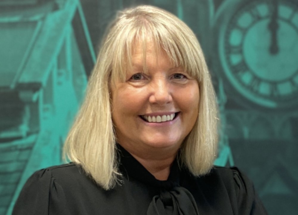 A person with shoulder‑length blonde hair wearing a black blouse stands in front of a teal backdrop featuring architectural illustrations and a large clock face.