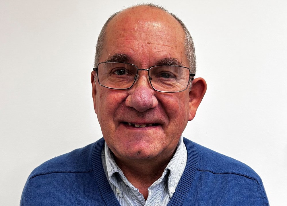 A person wearing a blue sweater over a light collared shirt sits against a plain white background, shown from the shoulders up.