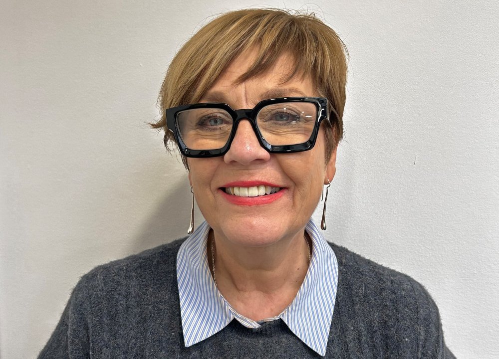 A person wearing a grey knit sweater layered over a blue‑striped collared shirt stands against a plain light‑coloured wall. Their short, light brown hair is neatly styled, and part of a pair of dark glasses is visible on one side.
